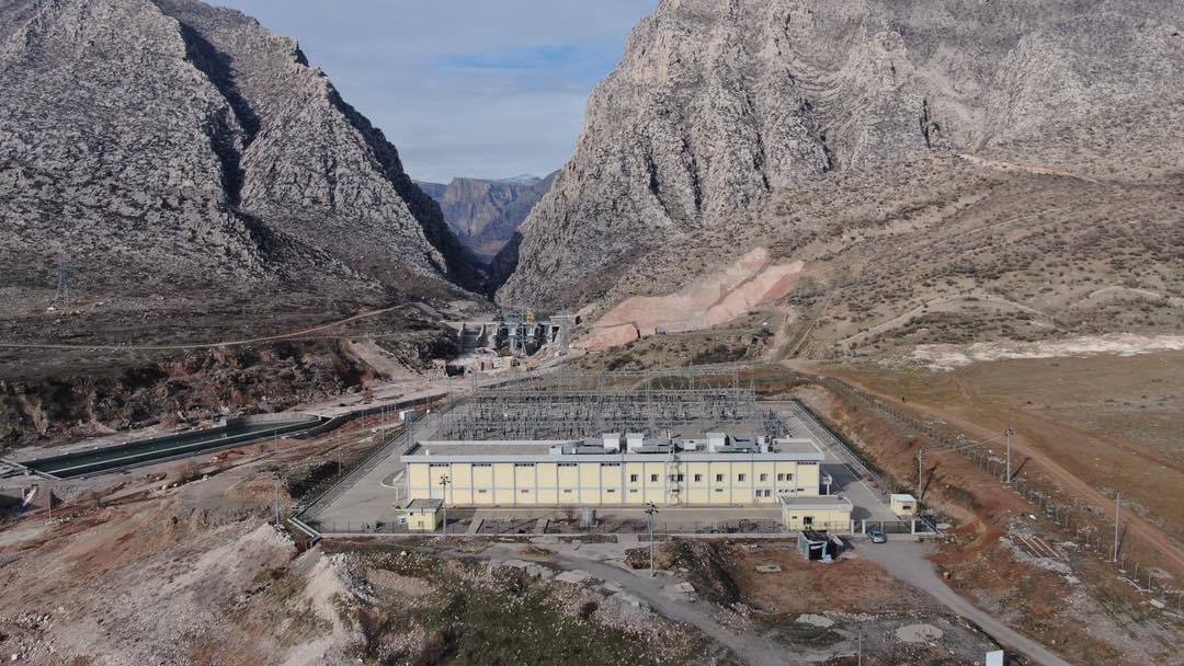 Electrical substation with a large yellow building set in a rocky mountain valley, surrounded by rugged terrain and infrastructure
